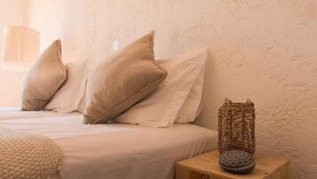 Una cama con ropa de cama y almohadas color beige, una cesta tejida con un objeto estampado sobre una mesita de noche de madera, y una pared con textura.