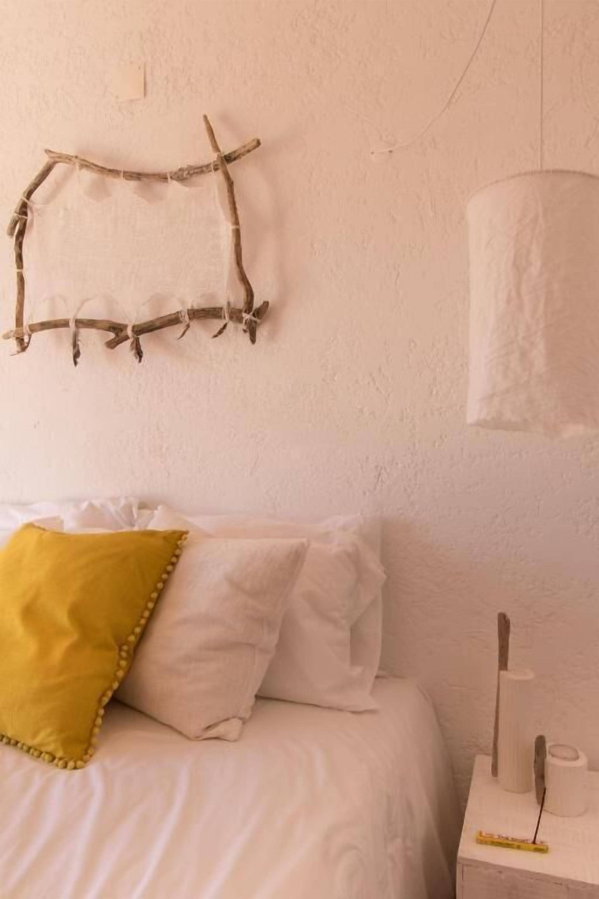 A bed with white bedding and a yellow pillow, a wall-mounted branch decoration, and a white nightstand with a lamp and a small object.