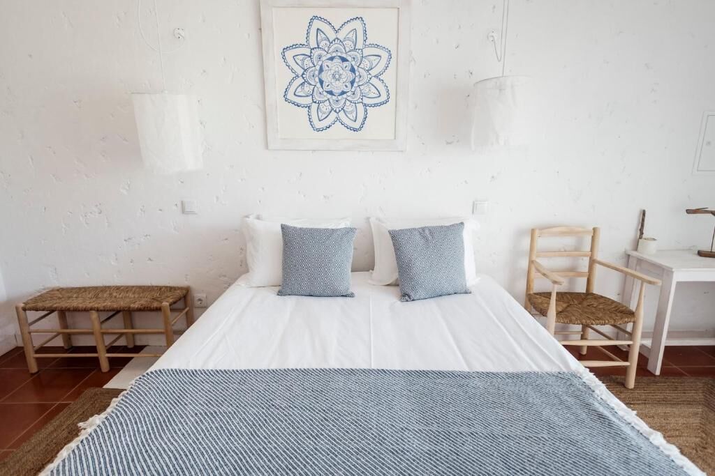 A bedroom with a bed, two pillows, a bedside table, a chair, a rug, and a wall art piece.