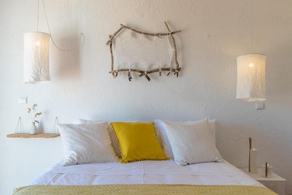 Een slaapkamer met een bed, witte en gele kussens, een hangende takdecoratie en twee wandlampen.