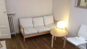 Une pièce avec un plancher en bois, un canapé blanc, une petite table ronde avec une lampe et une chaise blanche.