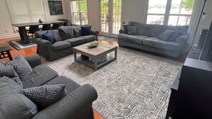 Ein geräumiges Wohnzimmer mit einem großen, grauen Ecksofa, einem Couchtisch und einem Deckenventilator. Große Fenster sorgen für Tageslicht und einen Blick ins Freie.