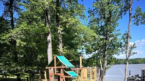 Ein Spielplatz mit Rutsche und Klettergerüsten, umgeben von Bäumen und Blick auf ein See.