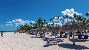 A beach with sun loungers and thatched umbrellas, palm trees, and people relaxing.