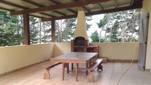 An outdoor patio with a wooden table and benches, a brick oven, and a view of trees.