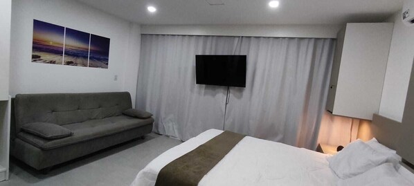 A bedroom with a bed, a sofa, a TV mounted on the wall, and a curtain.