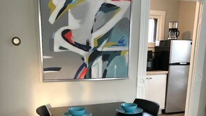 Een eettafel met twee blauwe kommen en stoelen, een modern abstract schilderij aan de muur, en een keuken op de achtergrond.