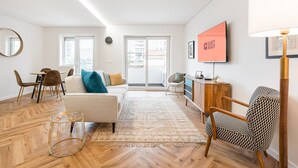En moderne stue med sofa, sofabord og en stue stol med mønster. Der er et fjernsyn på væggen og et vindue med udsigt til bygninger.