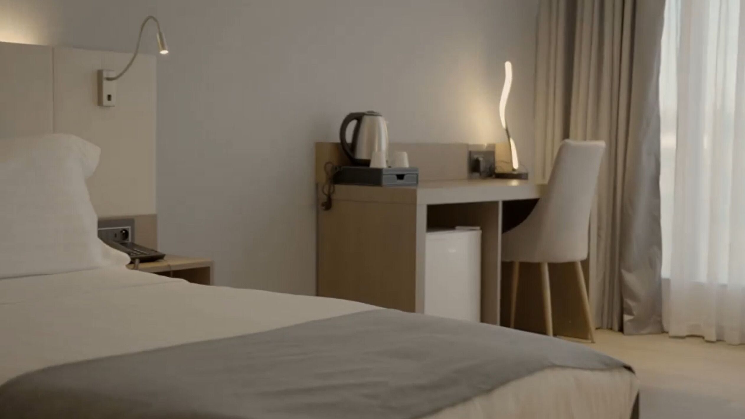 A hotel room with a bed, a desk with a kettle, a chair, and a nightstand.