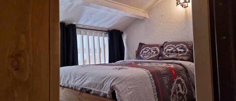 A bedroom with a wooden bed, patterned bedding, a window with curtains, and a wall-mounted light fixture.