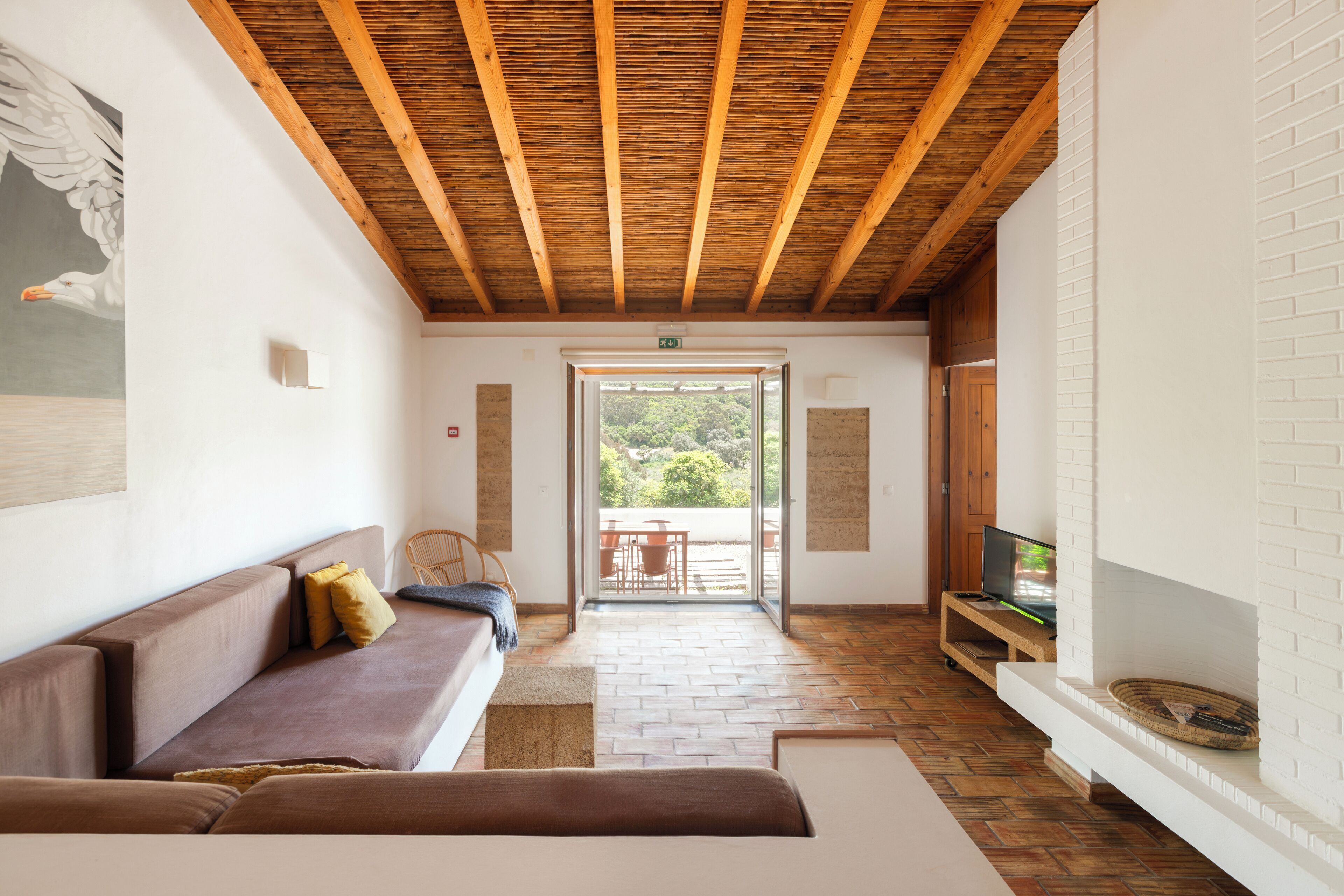 Un interno moderno con soffitto in legno, un divano marrone, un caminetto bianco e vista su una zona esterna attraverso una porta in vetro.