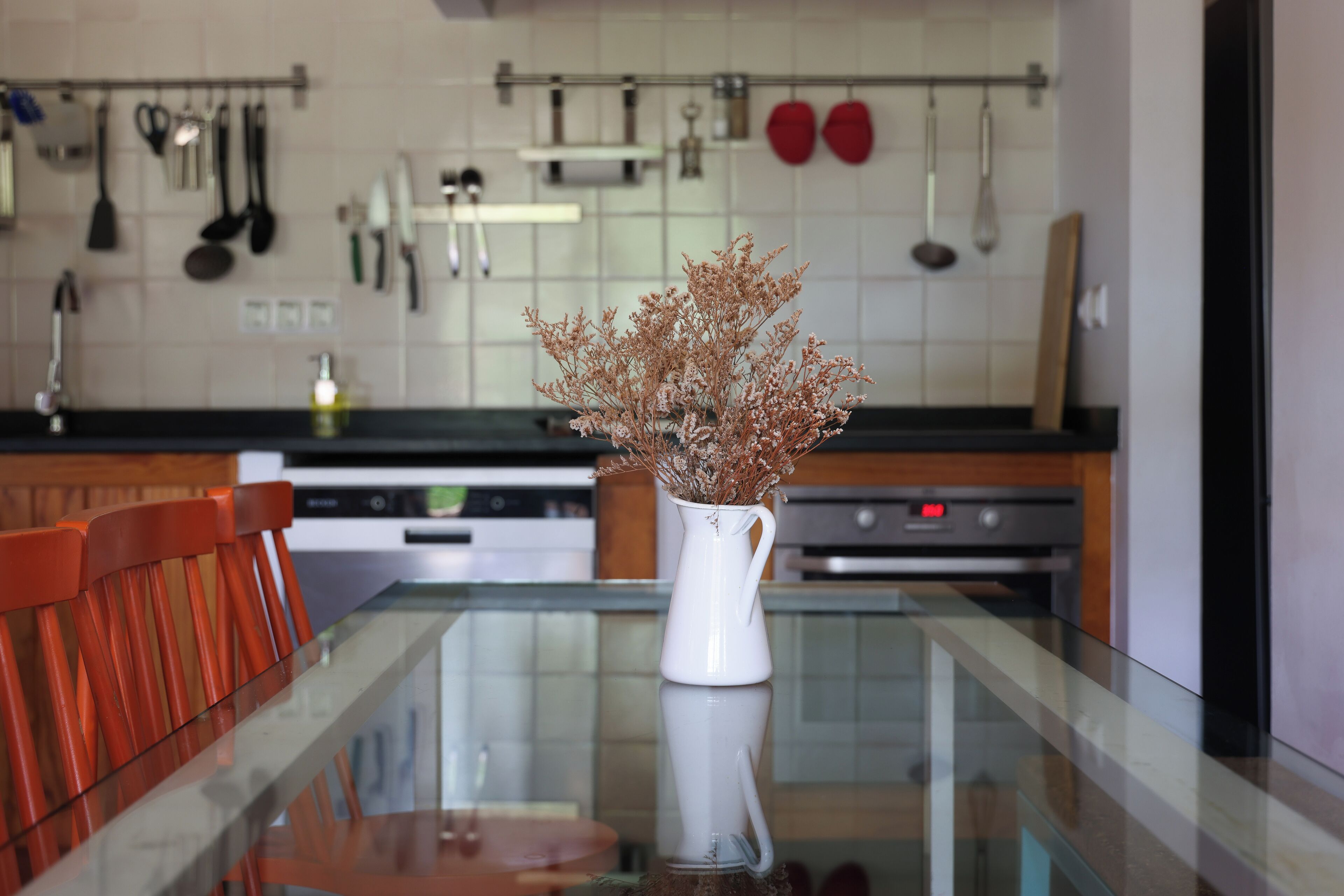 Una cocina con una mesa de vidrio, un jarrón blanco con flores secas y sillas de madera.