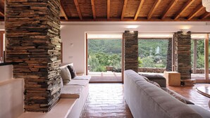 A modern living room with a stone pillar, a sofa, and a view of a lush landscape through large windows. - Monte da Vilarinha (Aljezur)