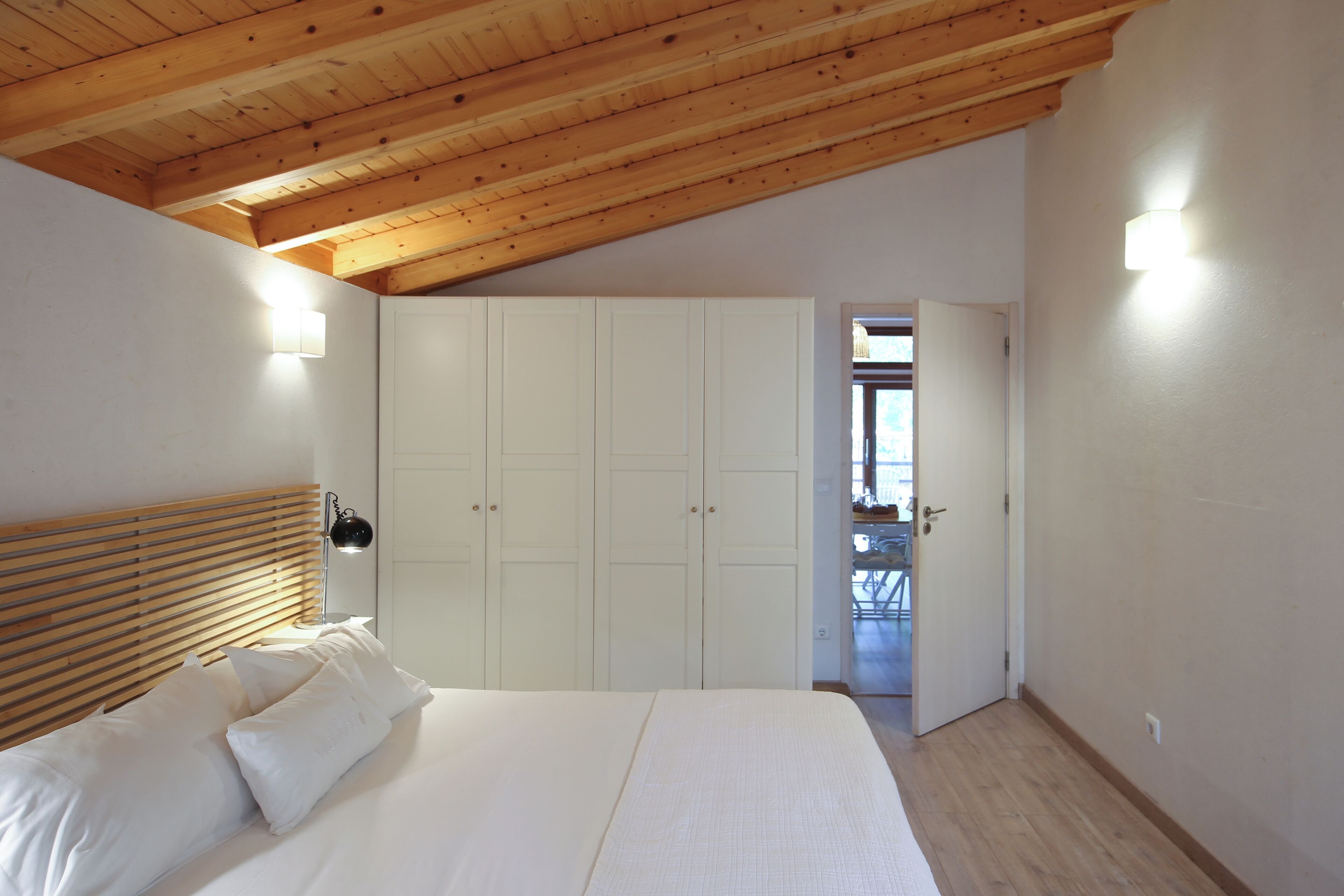 A bedroom with a wooden ceiling, a bed with white linens, a wardrobe, and a door leading to a balcony.