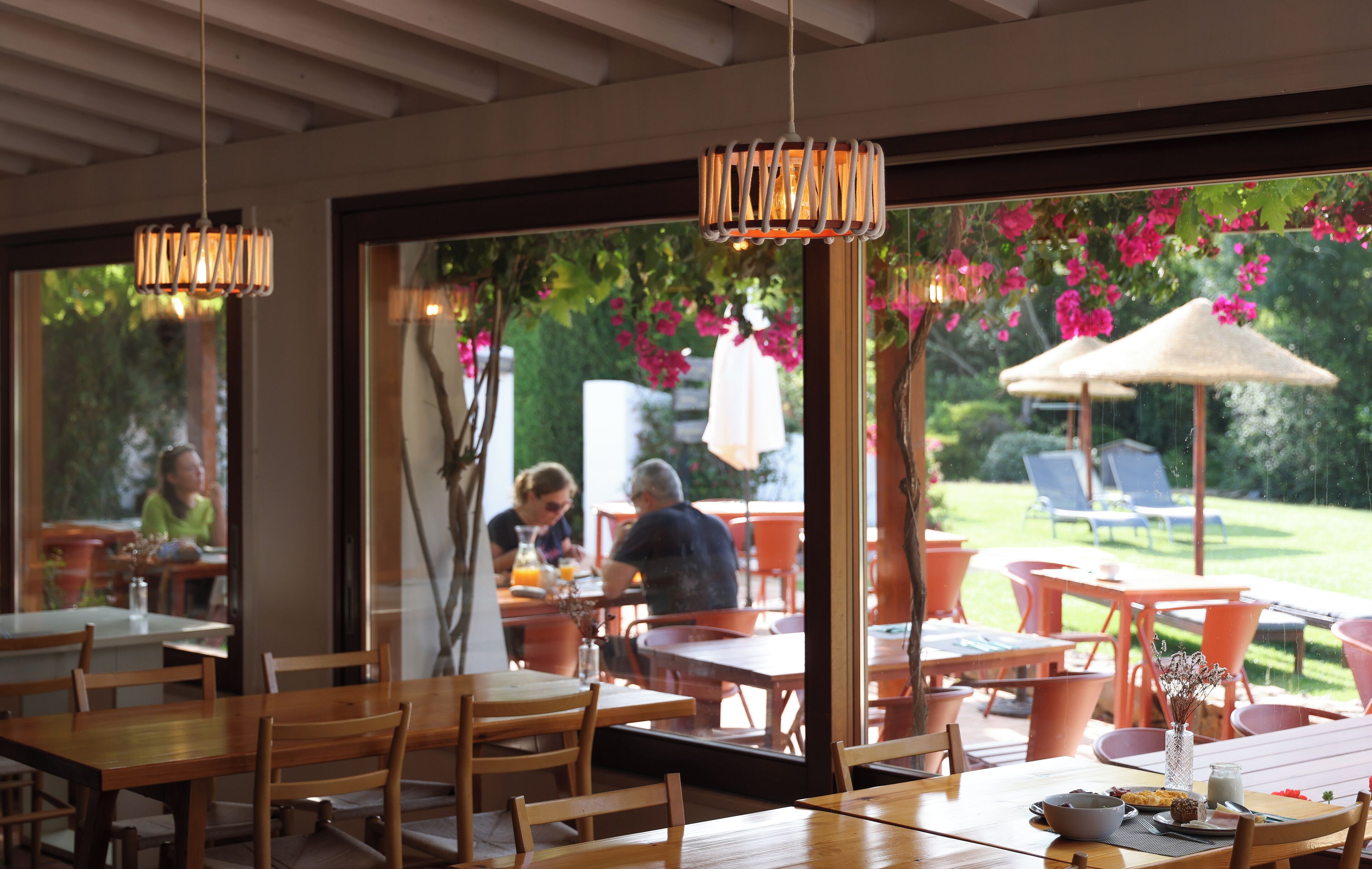 Un espace de restauration en plein air avec des tables et des chaises en bois, un luminaire suspendu et une vue sur un jardin agrémenté de fleurs roses et de parasols.