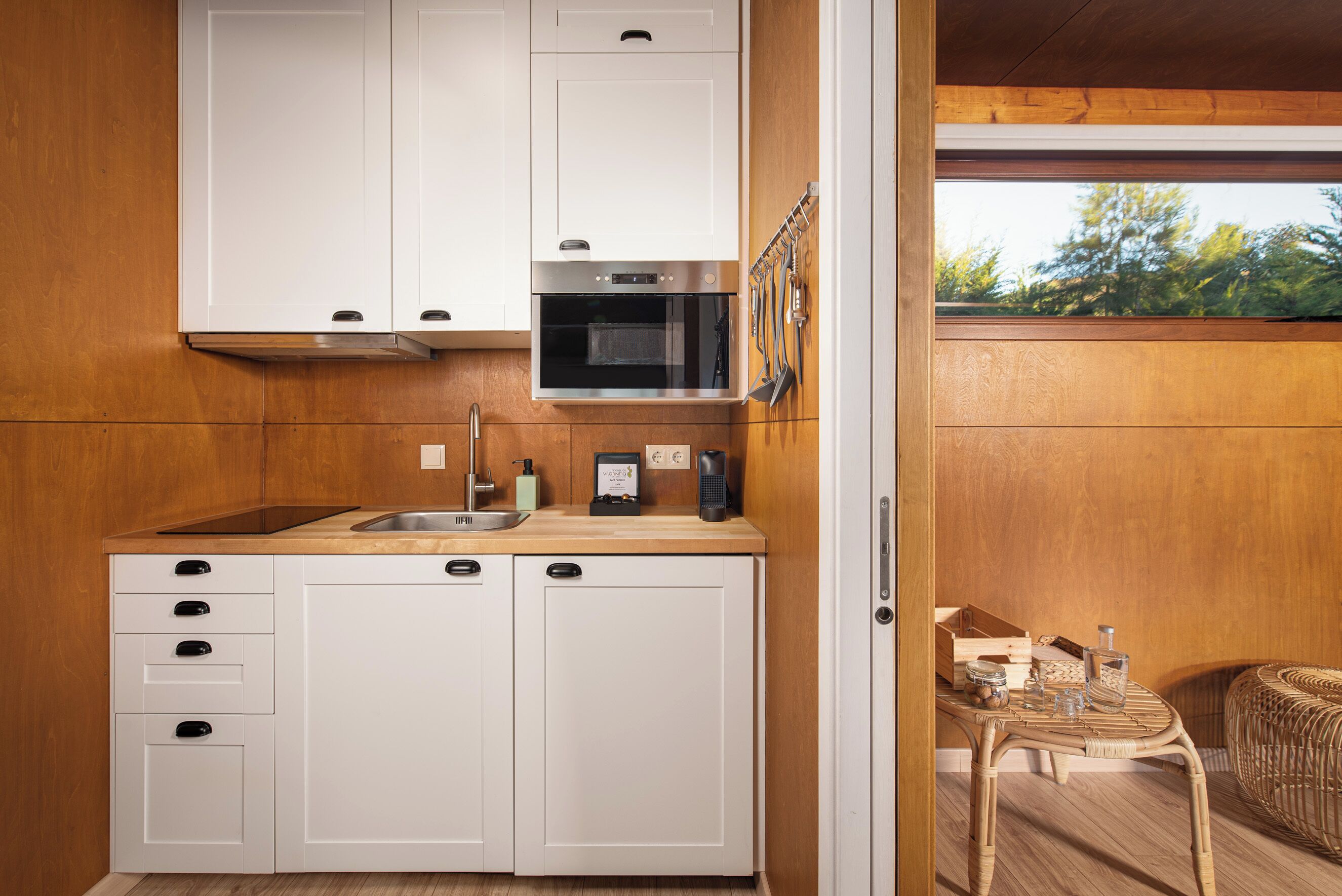 Cocina compacta con armarios blancos, microondas y un lavamanos. Se ve una silla de mimbre y una mesita con objetos encima.
