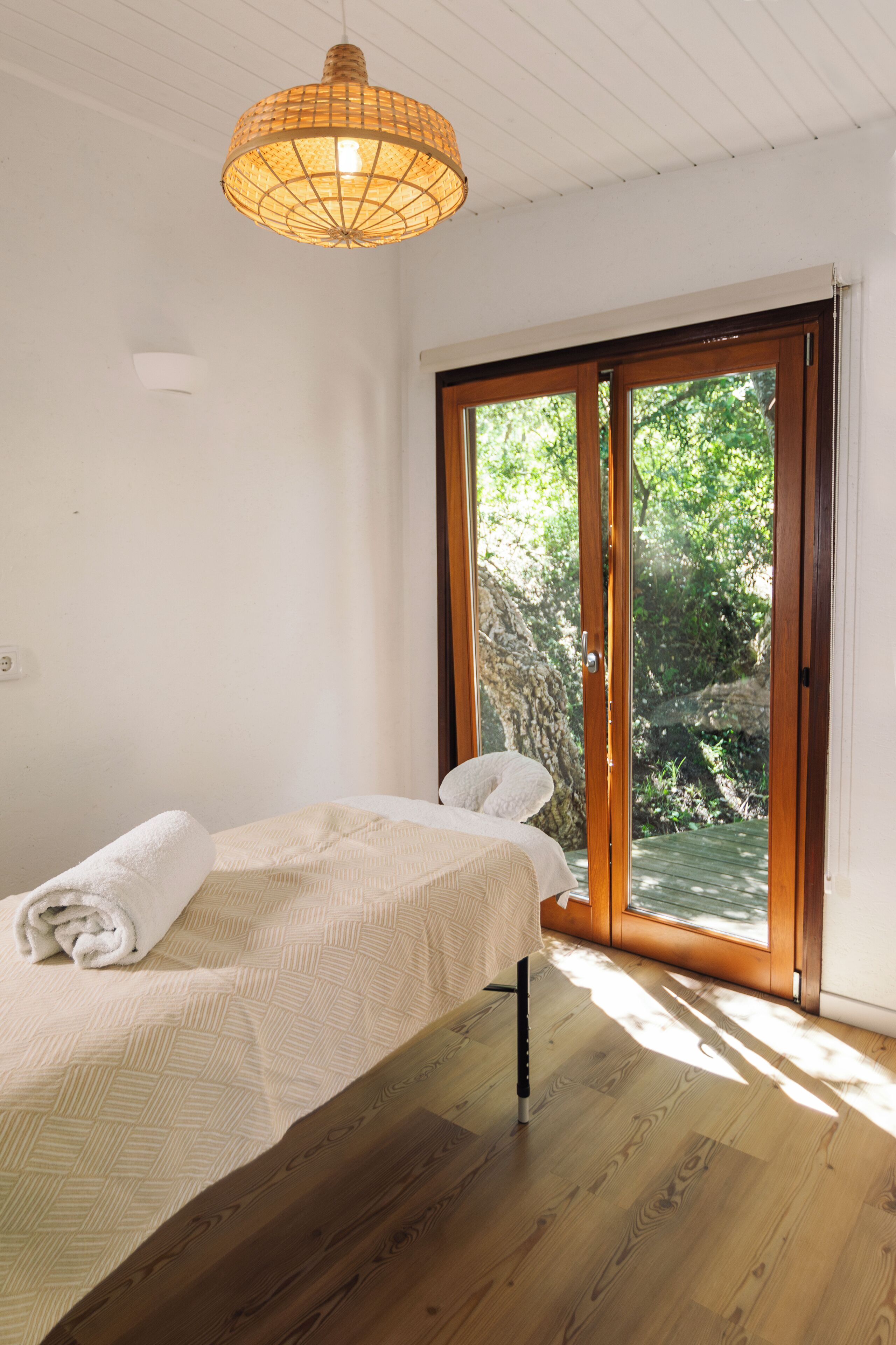 A bedroom with a bed, a wooden floor, a window with a view of greenery, and a hanging light fixture.