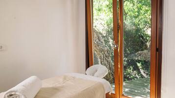 A bedroom with a bed, a wooden floor, a window with a view of greenery, and a hanging light fixture.