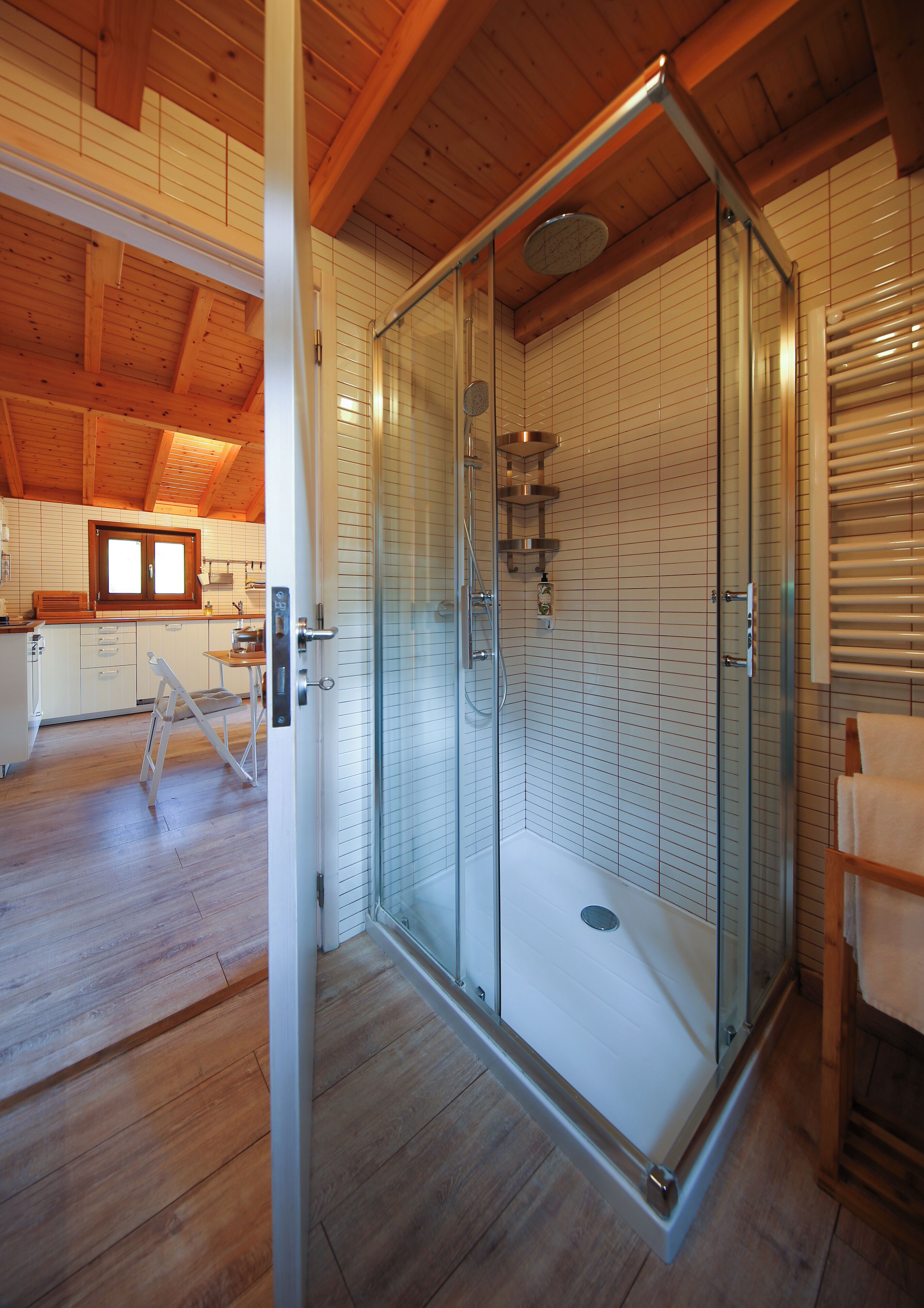 Ein modernes Badezimmer mit einer Duschkabine aus Glas, Holzboden und einem sichtbaren Küchenbereich im Hintergrund.