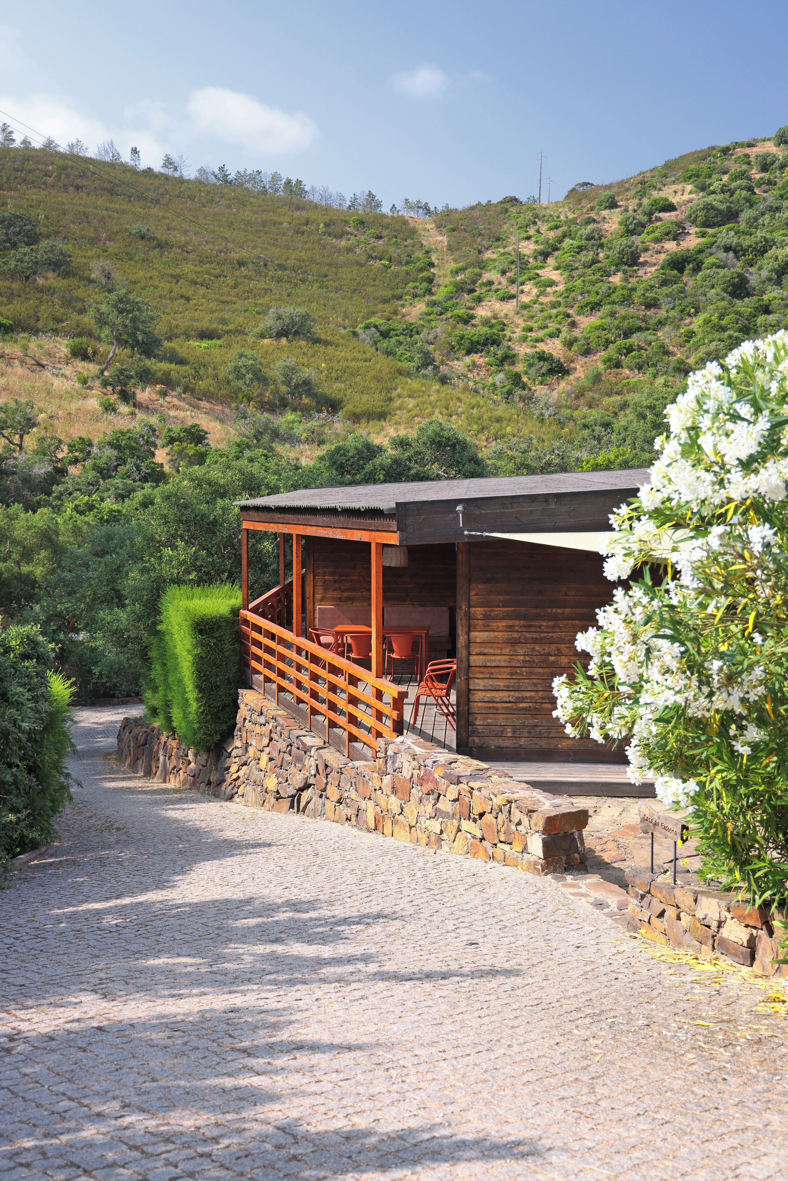 A rustic cabin with a stone wall entrance, a wooden deck with chairs, and a view of a hilly landscape.