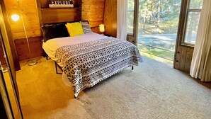 Un dormitorio de cabaña de madera con una cama, una mesita de noche, una lámpara y un ventanal con cortinas.