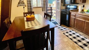 Un comedor con mesa y sillas de madera, mantel a cuadros y una cesta de limones. Se observa una cocina con estufa, microondas y armarios.