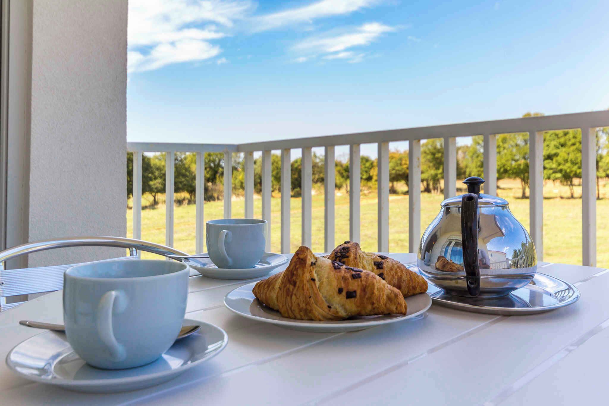 A table with a cup, a plate with croissants, and a coffee pot on a balcony with a view of trees.