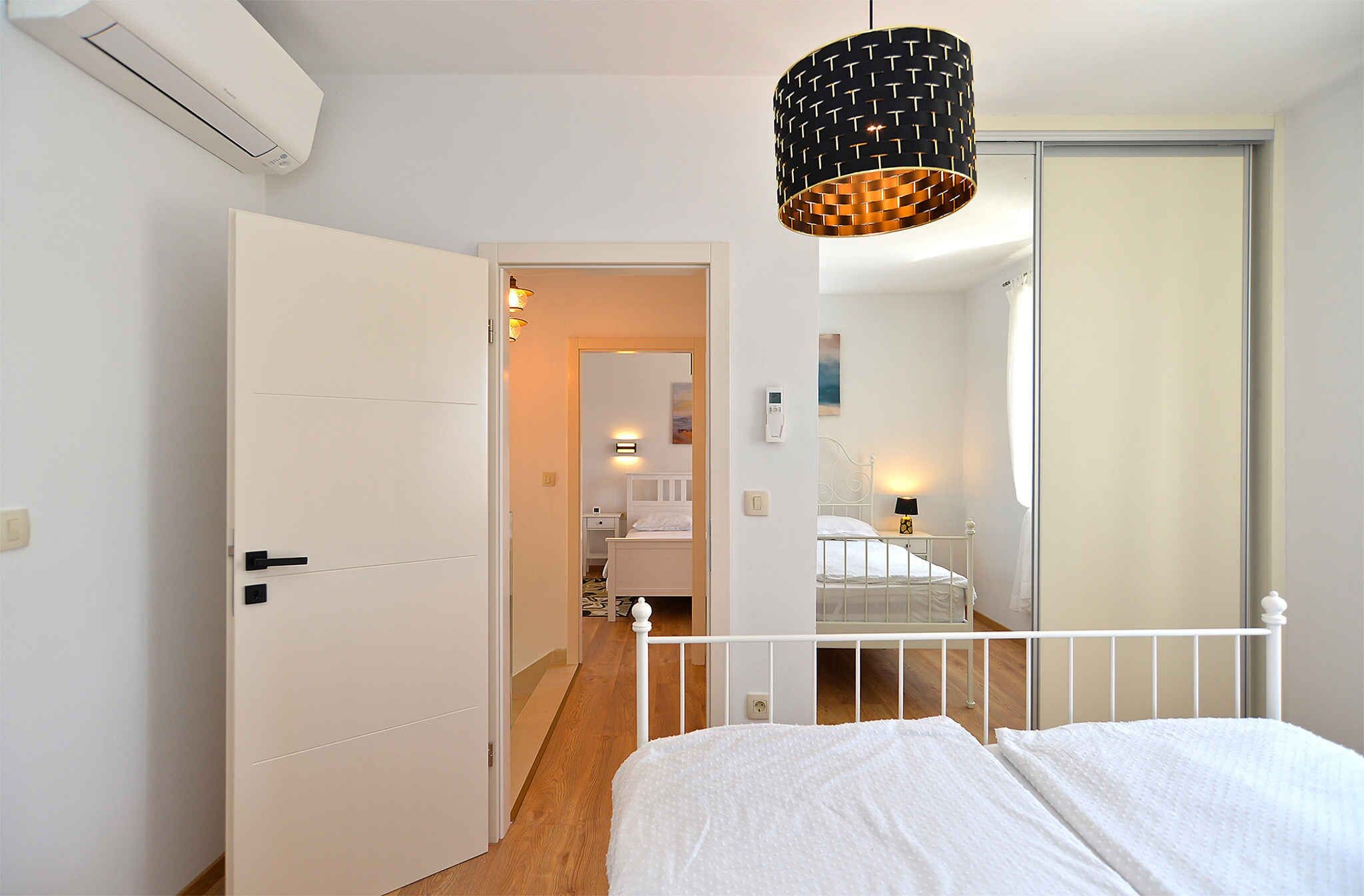 A bedroom with a bed, a white door, a hallway, a bathroom with a shower, and a hanging light fixture.