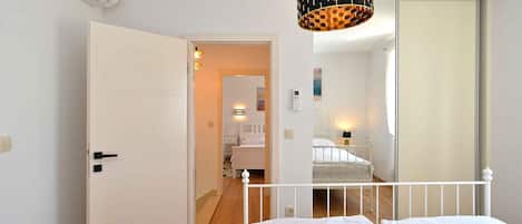 A bedroom with a bed, a white door, a hallway, a bathroom with a shower, and a hanging light fixture.