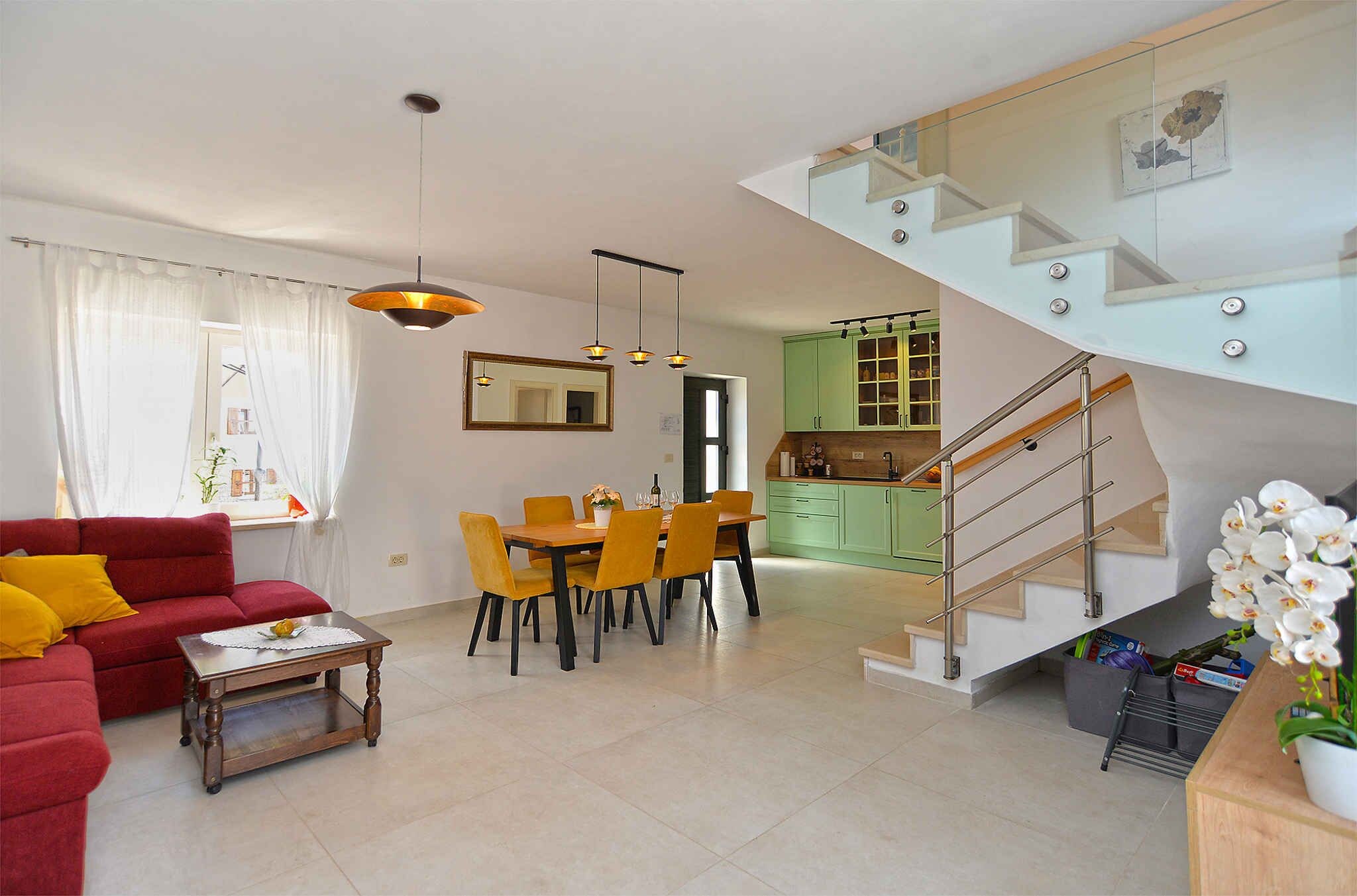 Ein modernes Wohnzimmer mit einem roten Sofa, gelben Sesseln, einem Holztisch und einer Essecke mit Tisch und Stühlen. Eine Treppe führt zu einer Küche mit grünen Schränken.