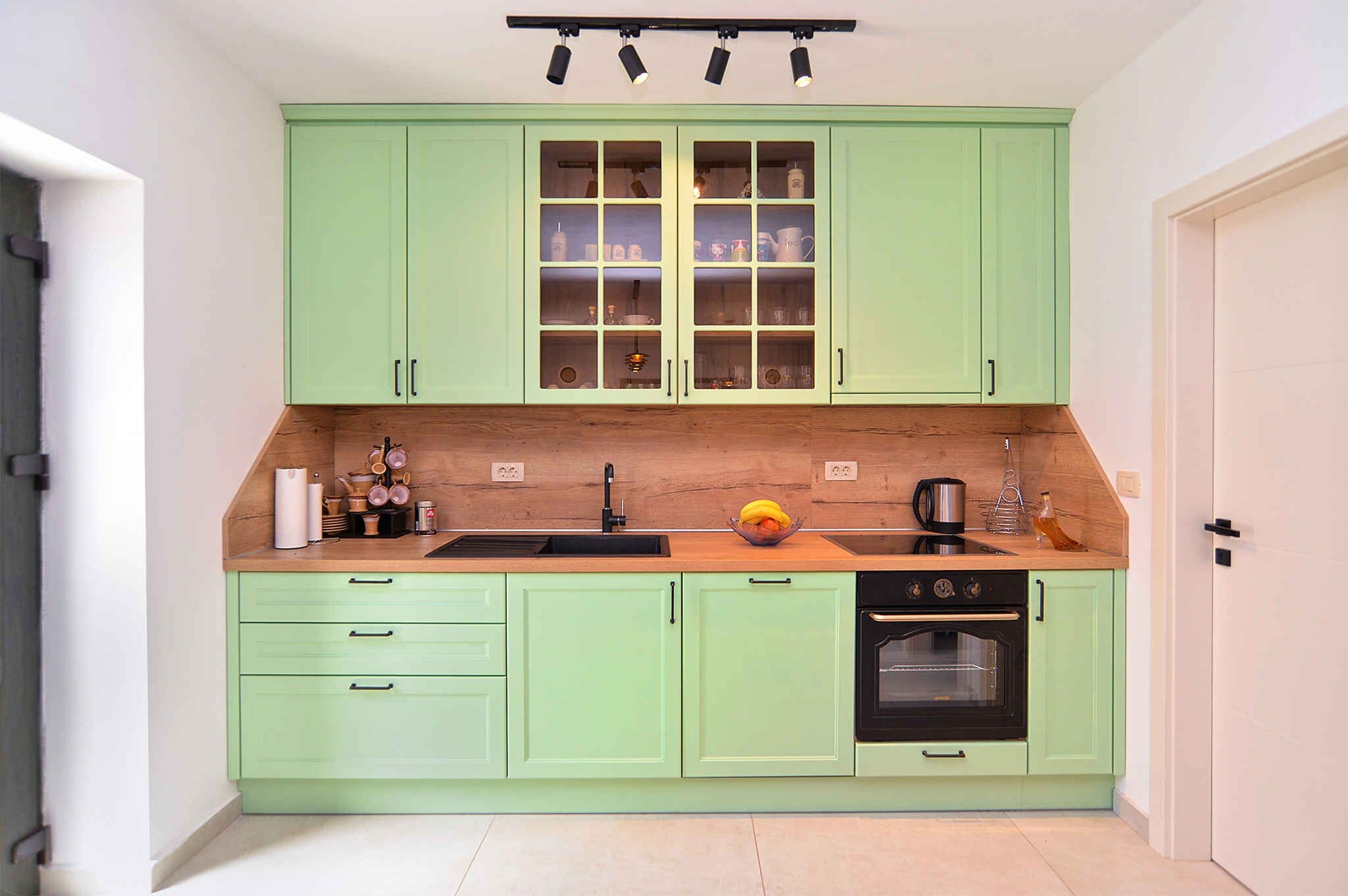 Une cuisine avec des meubles de couleur vert clair, un plan de travail en bois, un évier, une cuisinière et une fenêtre dotée de plusieurs étagères.