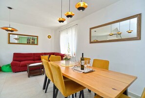 A dining area with a wooden table, yellow chairs, and a red sofa. There are hanging pendant lights, a large mirror, and a window with curtains.