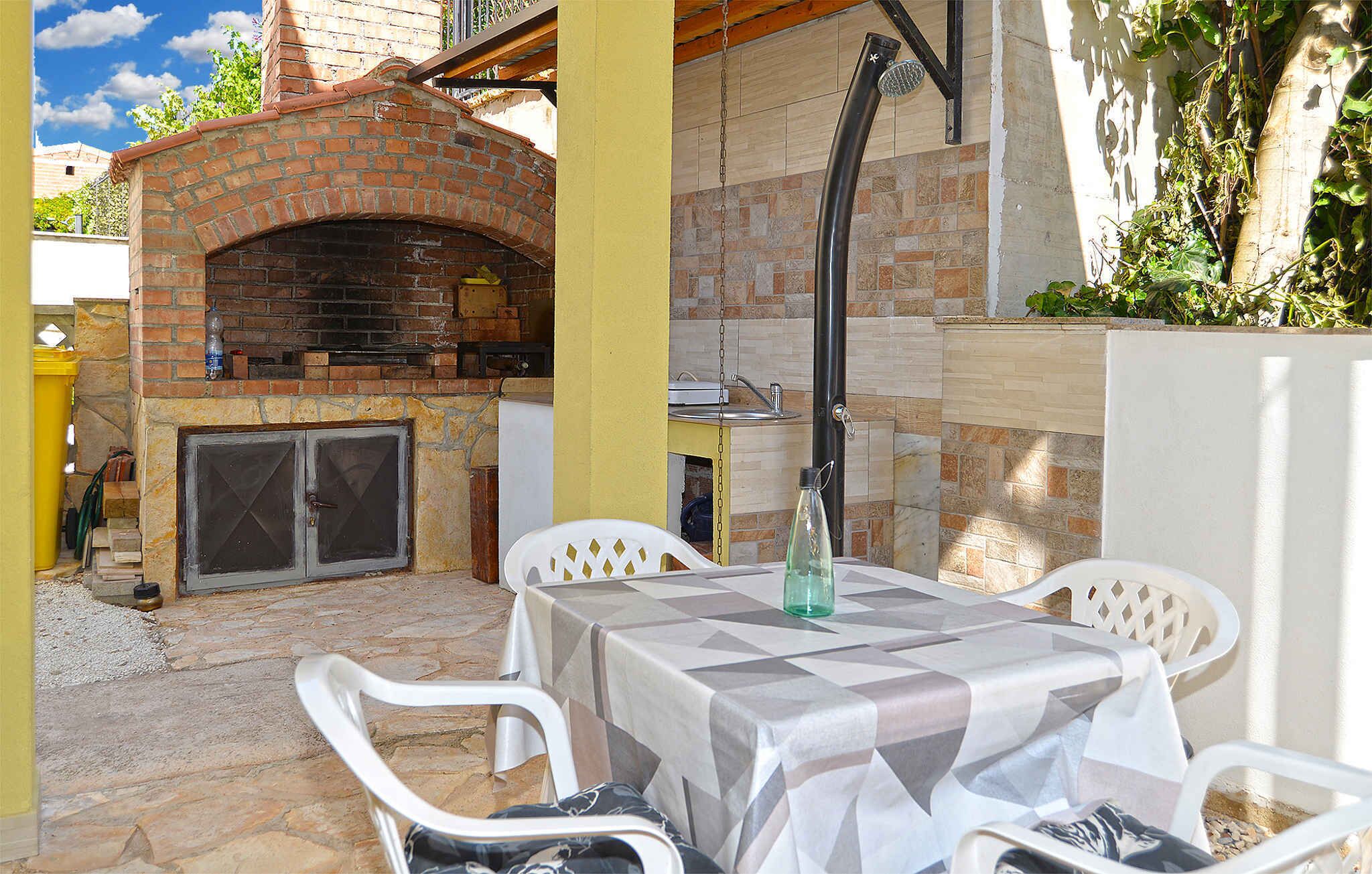 Eine Terrasse im Freien mit einem Backofen aus Ziegeln, weißen Plastikstühlen und einem Tisch mit kariertem Tischläufer.