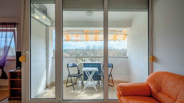 A room with a sliding glass door leading to a balcony with a table and chairs, a sofa, and a view of trees. - Holiday apartment mit Klimaanlage und Balkon (Monterol)