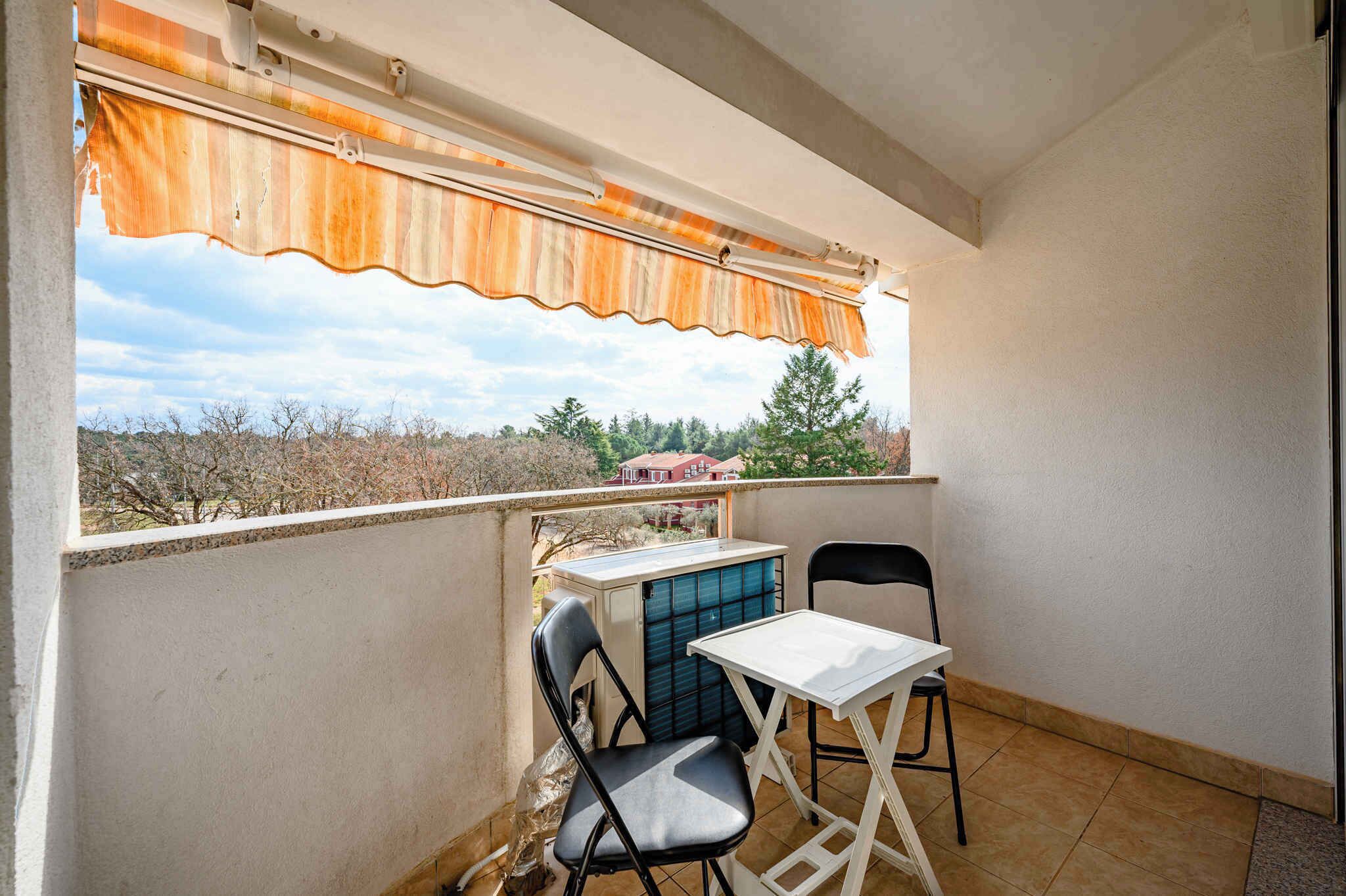 A balcony with a table and chairs, a window with a striped awning, and a view of trees and buildings.