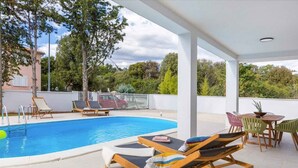 Área de piscina com espreguiçadeiras, mesa e vista para árvores e edifícios.