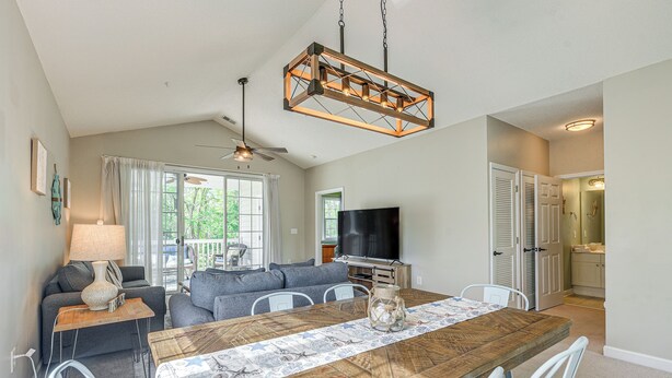 Un coin repas avec une table en bois, des chaises blanches, un ventilateur de plafond et un téléviseur en arrière-plan.