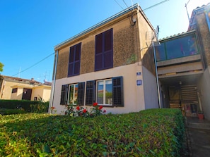 Un bâtiment à deux étages, doté d’une façade beige, de volets sombres et d’un petit jardin agrémenté de fleurs rouges.
