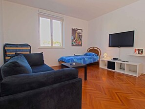 Une pièce comprenant un canapé, un lit avec une couverture bleue, un téléviseur posé sur un meuble et une fenêtre avec des stores.