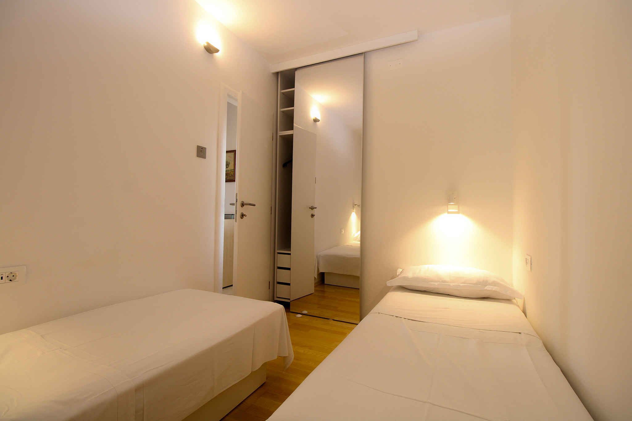 Una habitación pequeña y bien iluminada con dos camas individuales, suelo de madera y un armario con puertas correderas.