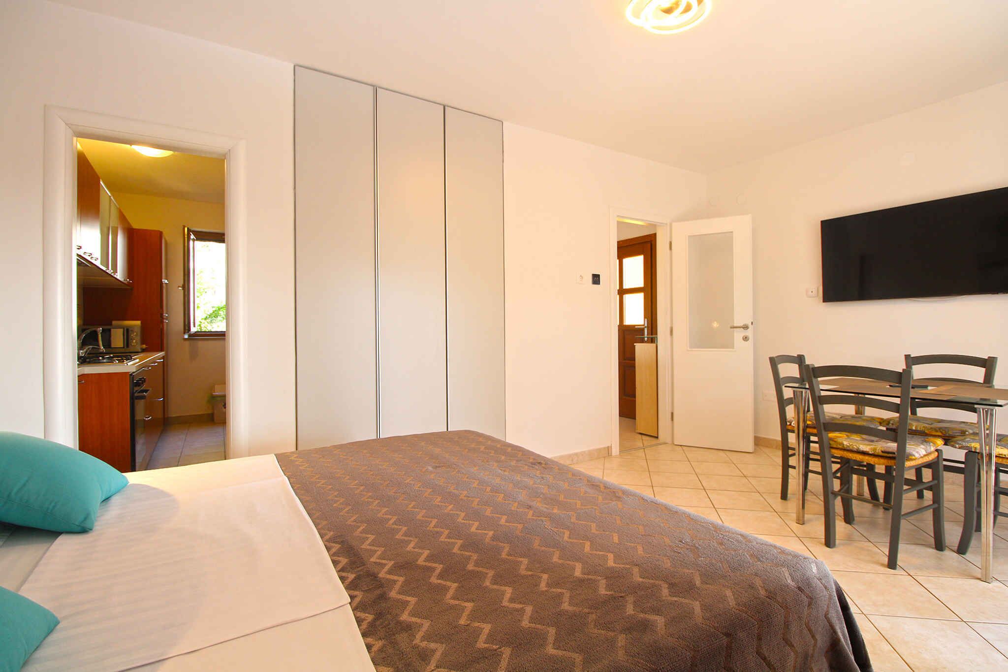 A bedroom with a bed, a dining table with chairs, a TV mounted on the wall, and a kitchen area visible through an open door.