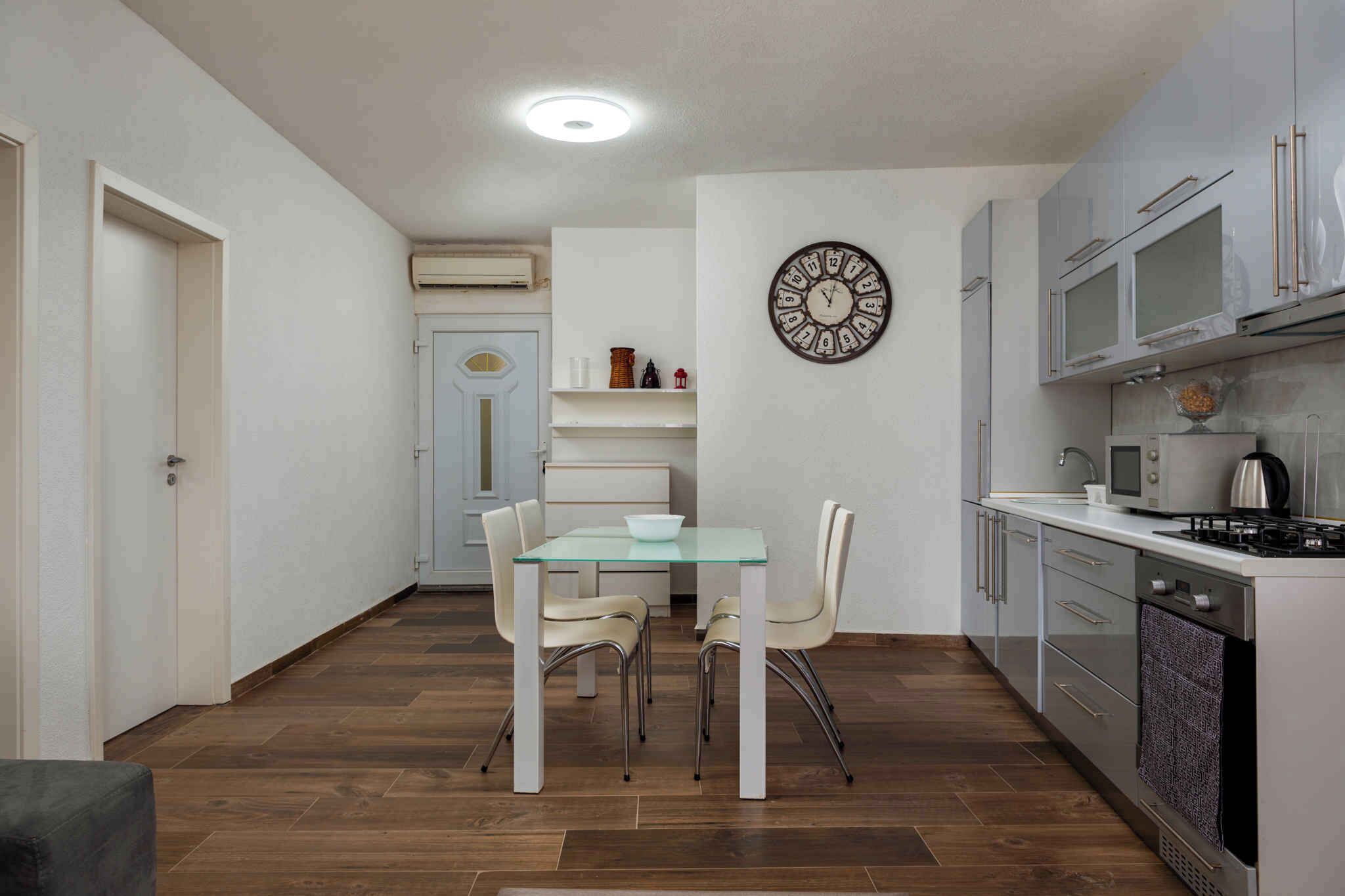 A compact kitchen with a dining area, featuring a glass table, white chairs, a wall clock, and modern appliances.