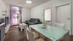 A modern living room with a glass dining table, grey sofa, white chairs, and a TV. - Holiday apartment with air conditioning (Pula)