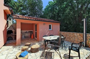 Um pátio externo com piso de pedra, uma mesa com cadeiras, uma pequena área de cozinha e uma parede de pedra.