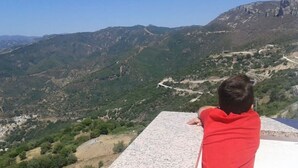 A person in a red shirt standing on a stone platform overlooking a mountainous landscape with a winding road. - Mountain Relaxation  (Baunei)