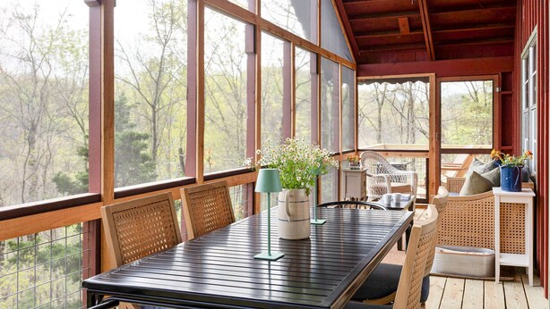 Een houten terras met een tafel, stoelen, een plafondventilator en uitzicht op bomen door grote ramen.