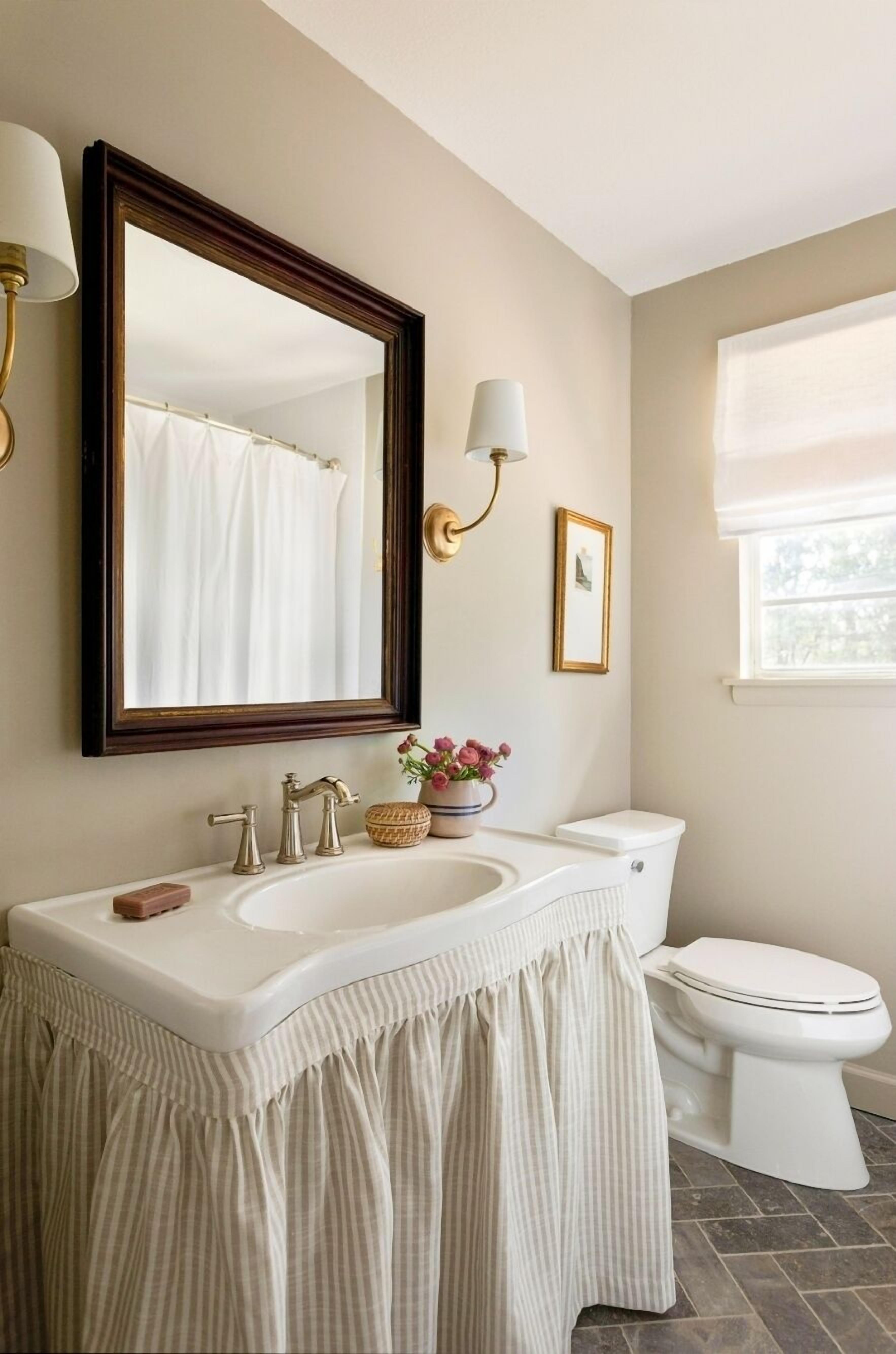 Une salle de bain avec un grand miroir, un lavabo à deux robinets, des toilettes et une fenêtre avec un rideau.