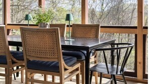 Een overdekt terras met houten tafels en stoelen, grote ramen en uitzicht op bomen.