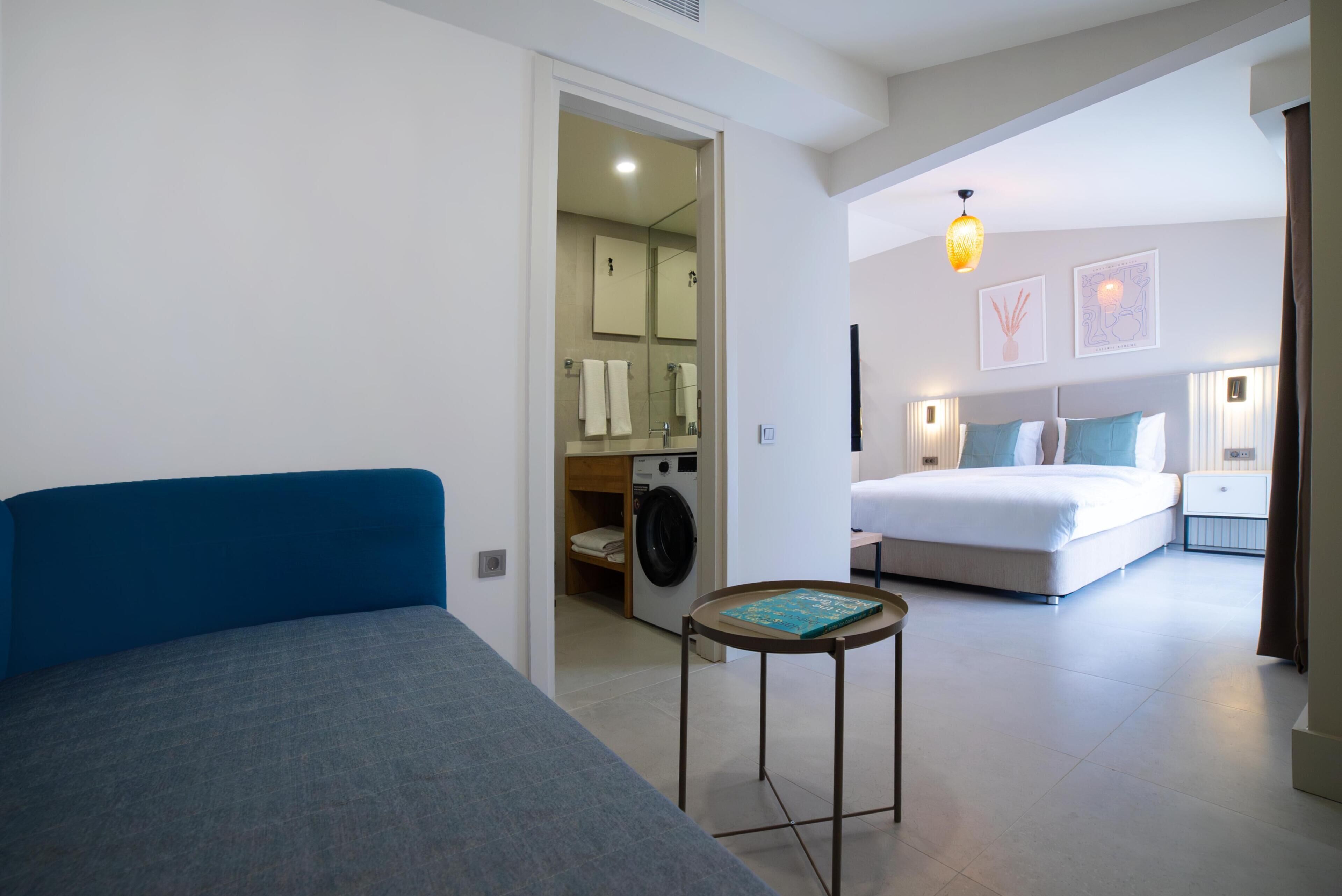 A modern hotel room with a blue sofa, a small round table, a bed with white bedding, a washing machine, and a mirror.