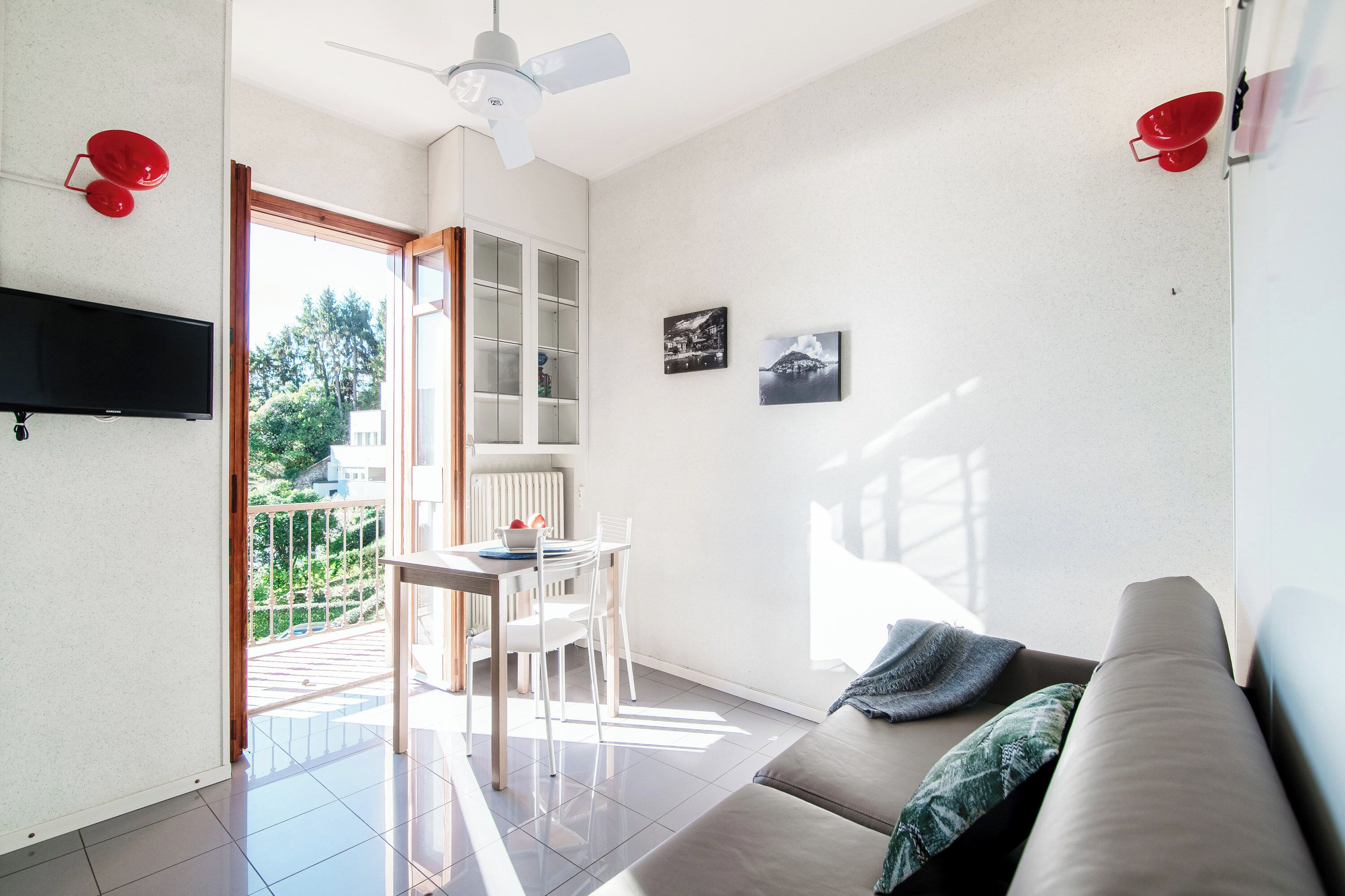 A modern living room with a sofa, a dining table with chairs, a TV mounted on the wall, and a balcony with a view of greenery.
